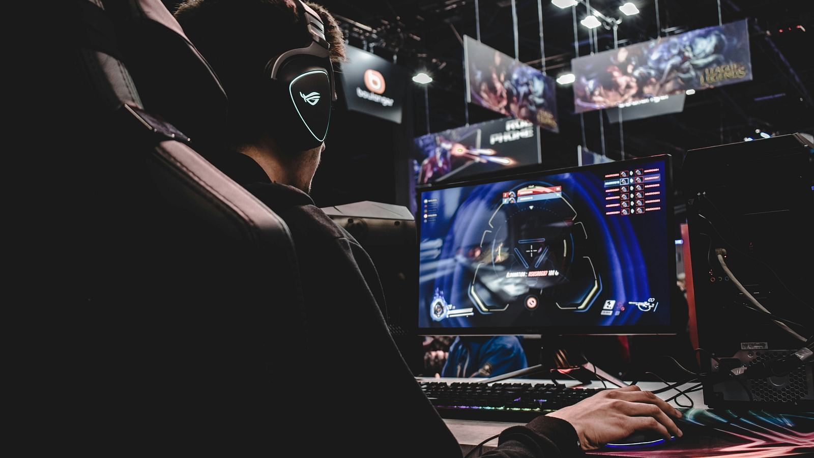 Side view from the back of a man wearing gaming equipment and playing games on a machine. There are screens in the background.