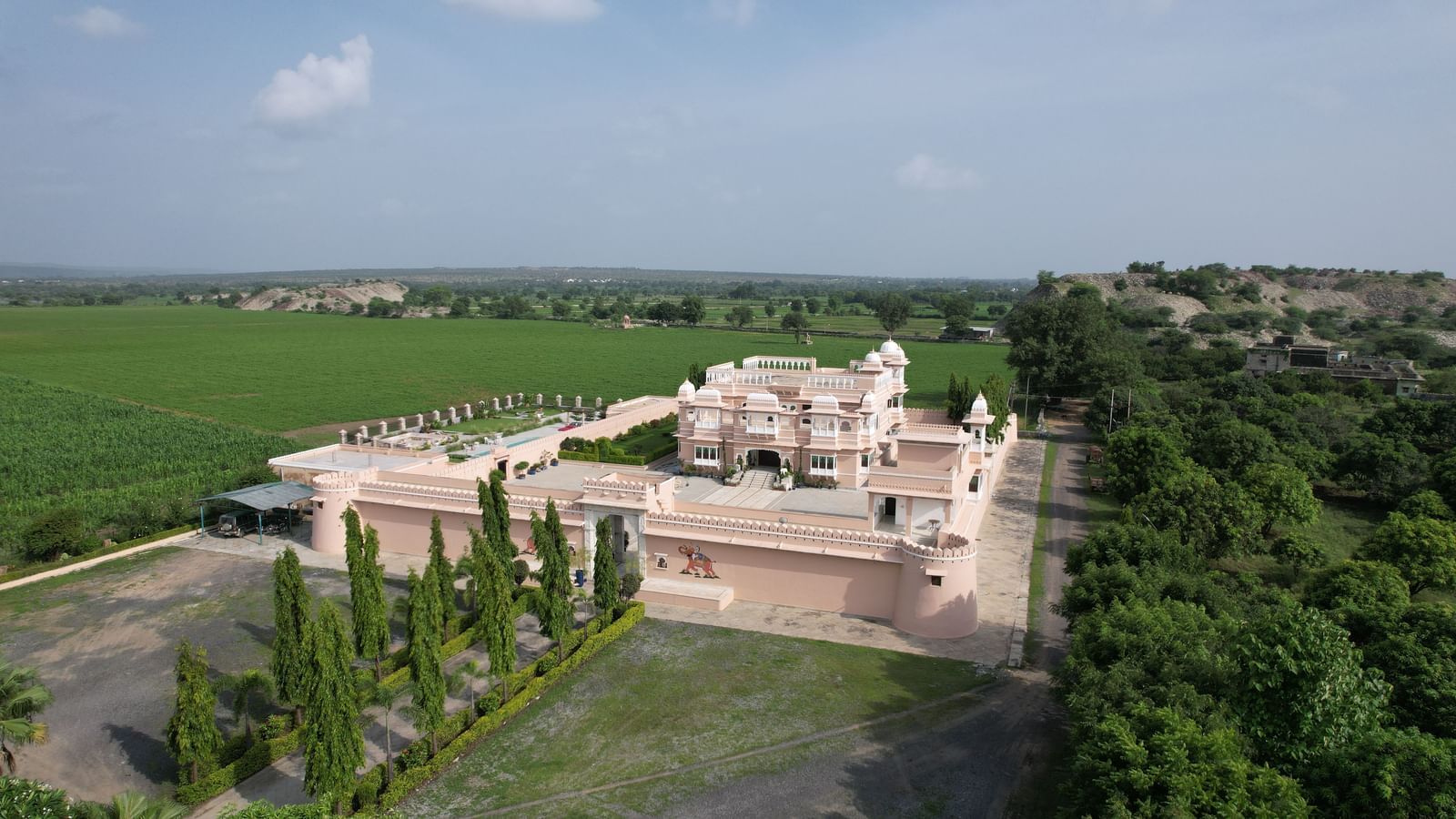 An aerial view of a large, light-colored building complex surrounded by green trees and fields at Mahendra Niwas. A paved drive and a landscaped garden are visible in the foreground.