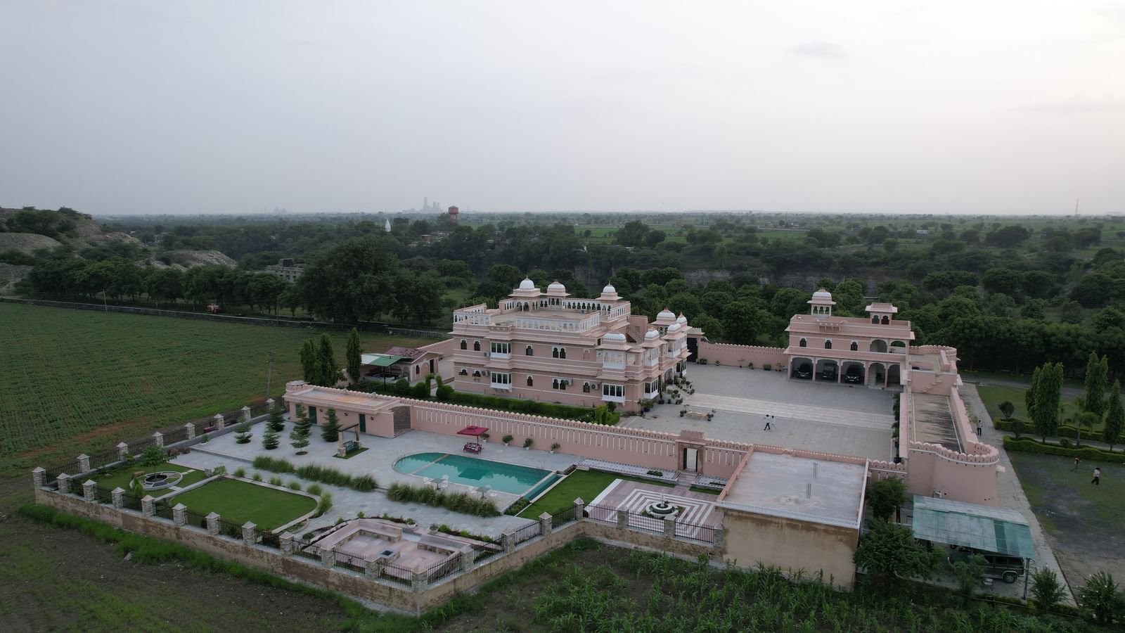 A high-angle aerial view of Mahendra Niwas, presenting the entire property with its intricate garden design, swimming pool, and surrounding landscape, all under a clear sky.
