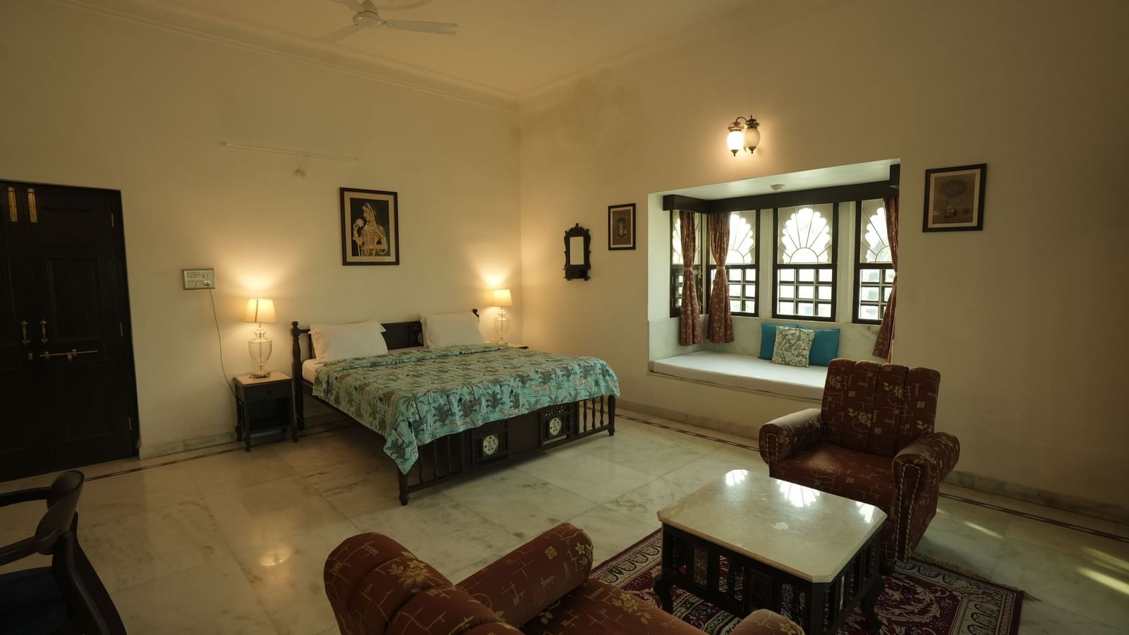 A wide-angle view of the Heritage Courtyard Room at Mahendra Niwas, featuring a large bed, a plush seating area, and traditional windows with curtains.