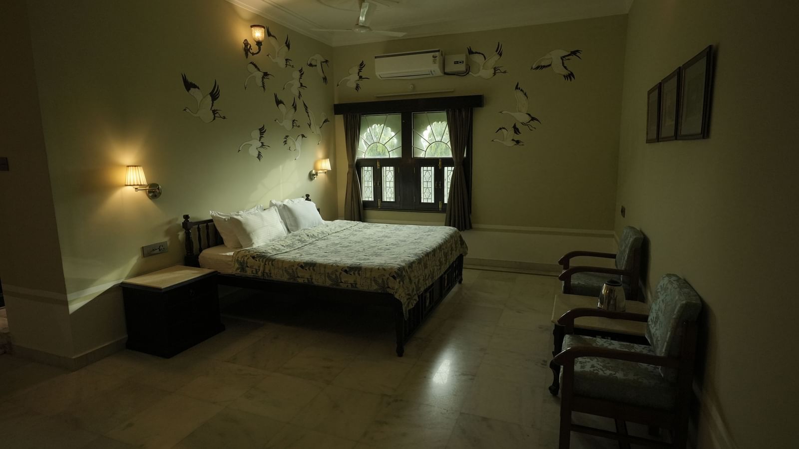 A wide, low-angle shot of the Deluxe Room at Mahendra Niwas. A bed with a light-colored headboard is pushed against a wall decorated with a pattern of black, stylized birds.