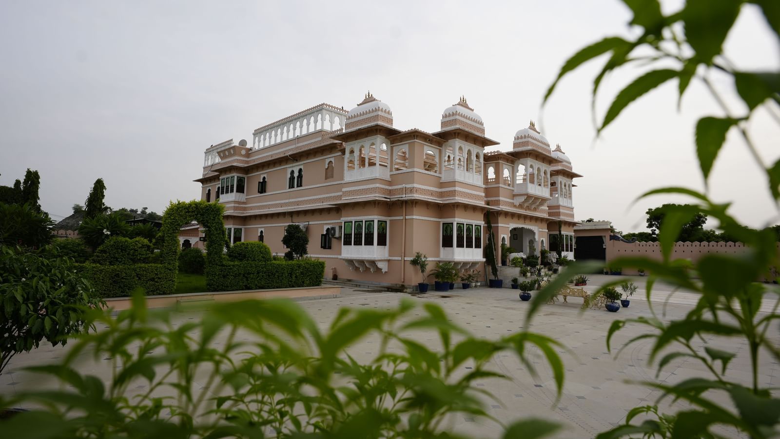 A low-angle exterior view of Mahendra Niwas, framed by vibrant green foliage, giving a sense of the property's elegant architecture nestled within a beautiful garden.