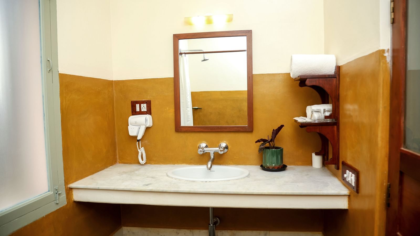 A bathroom with a sink, toiletries, and a mirror on the wall, A sitting area, a cupboard, and a tall lamp | Mango Hill La Serene, Pondicherry
