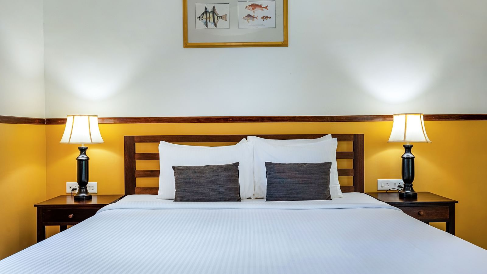 A neatly made double bed with pillows, flanked by matching lamps on nightstands, beneath a framed picture in a room near Pondicherry | Mango Hill Shallow Beach Resort ECR