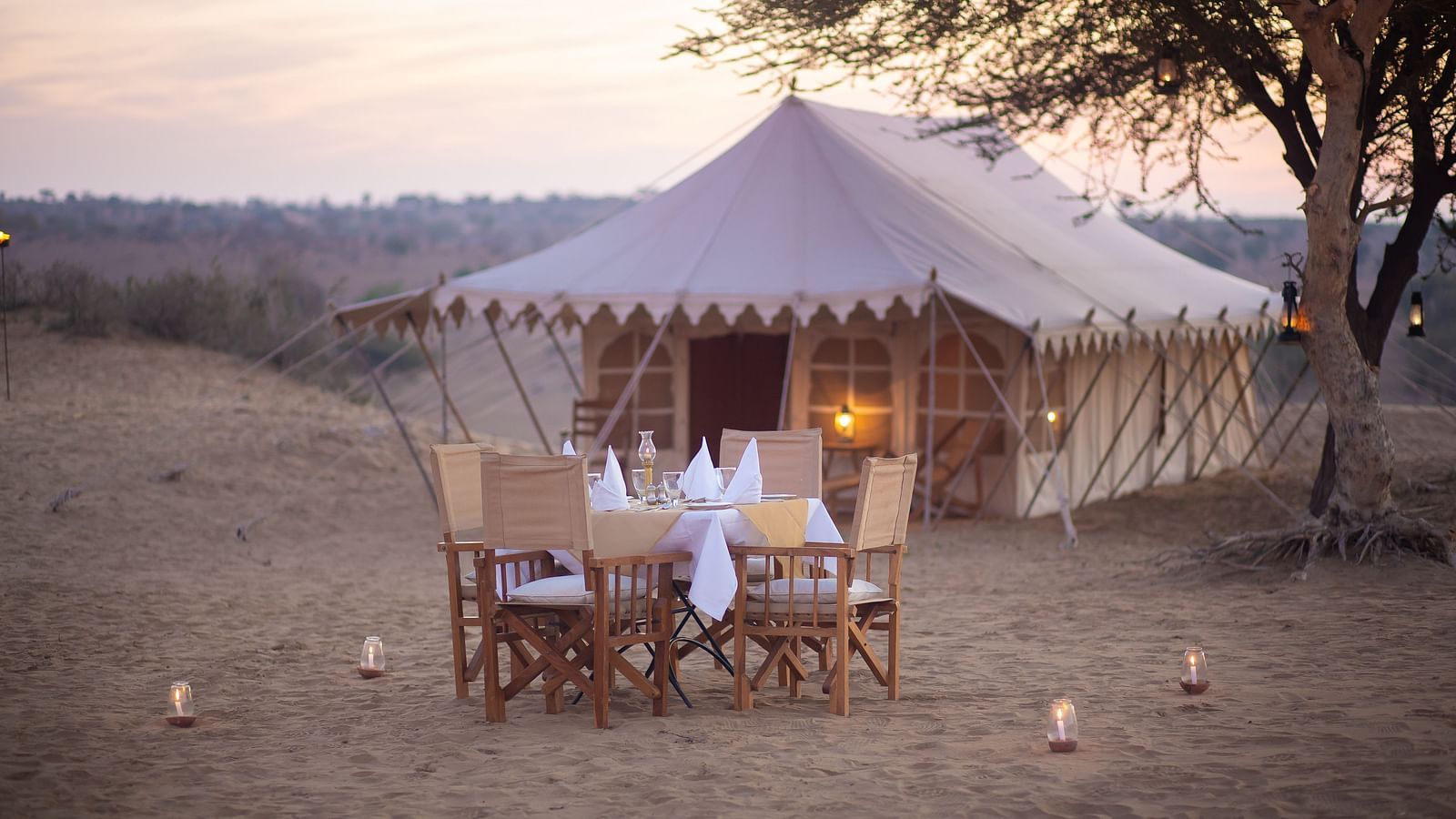 An outdoor dining set awaits by a large tent in the desert, shaded by a tree, for an intimate meal amidst the tranquility of nature.- at Manvar Resort 