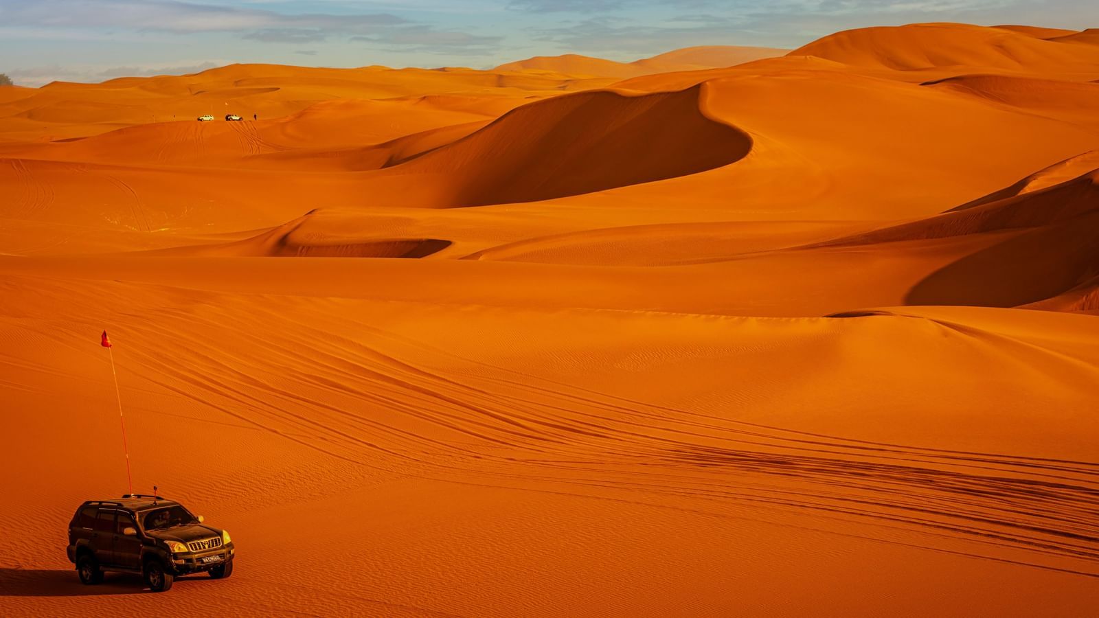 A four-wheel-drive vehicle navigates the steep, orange sand dunes of a desert landscape under a blue sky.