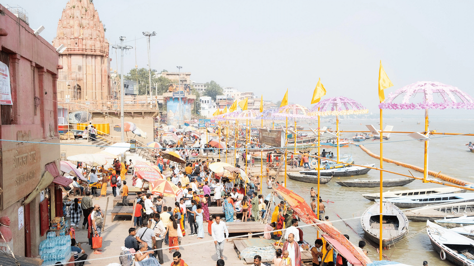 Crowds gathered along the riverbank near temples and boats at a ghat in India.