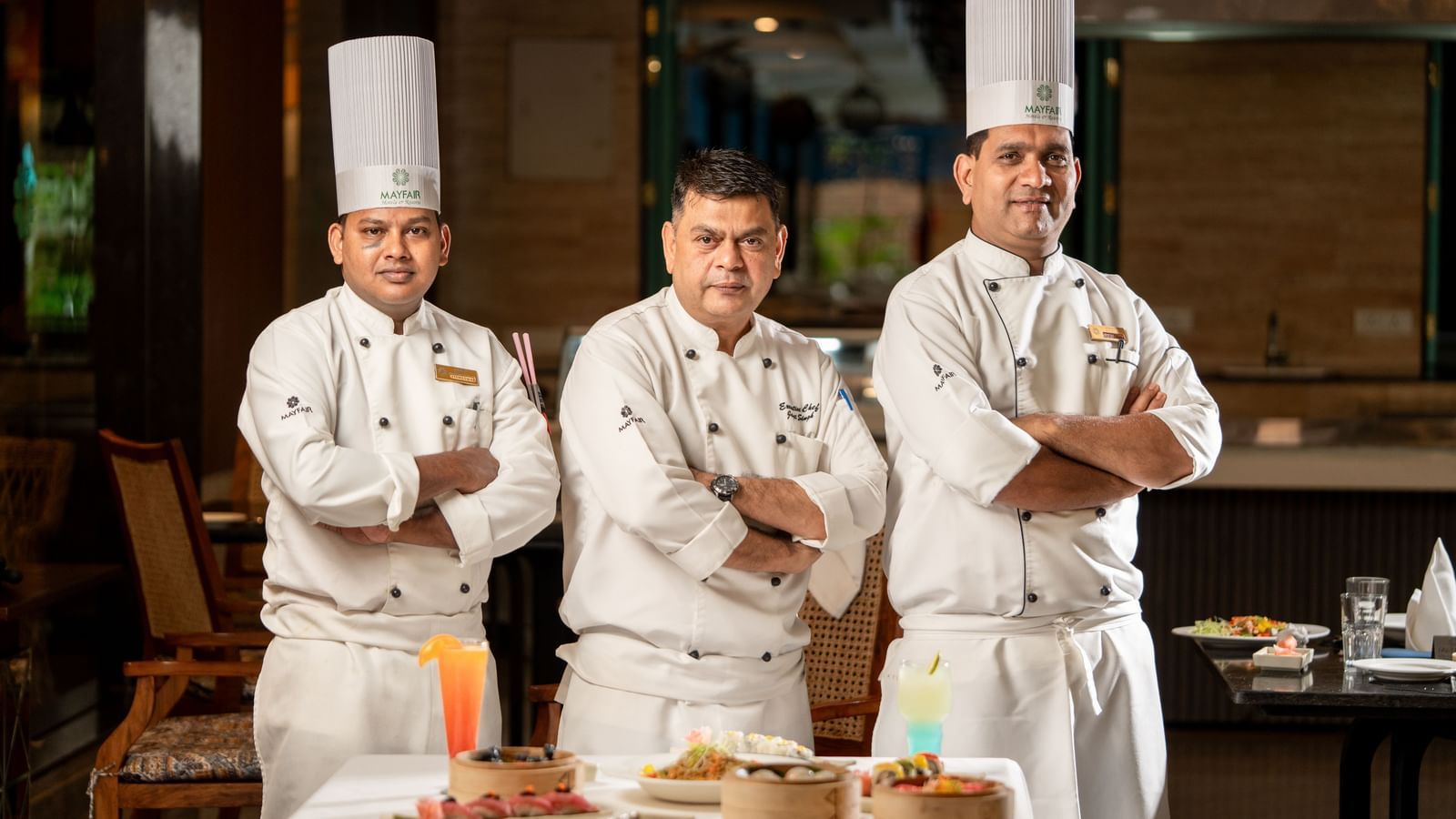 Three chefs in white uniforms standing confidently with folded arms behind a table set with dim sum, desserts, and drinks in a restaurant setting.