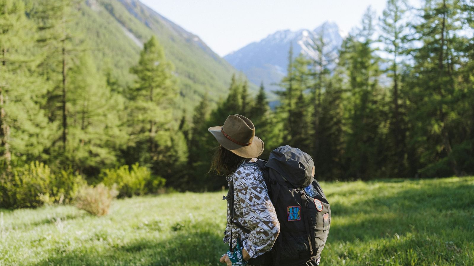  A traveller carrying a backpack walking through a green valley surrounded by tall trees and mountains.
