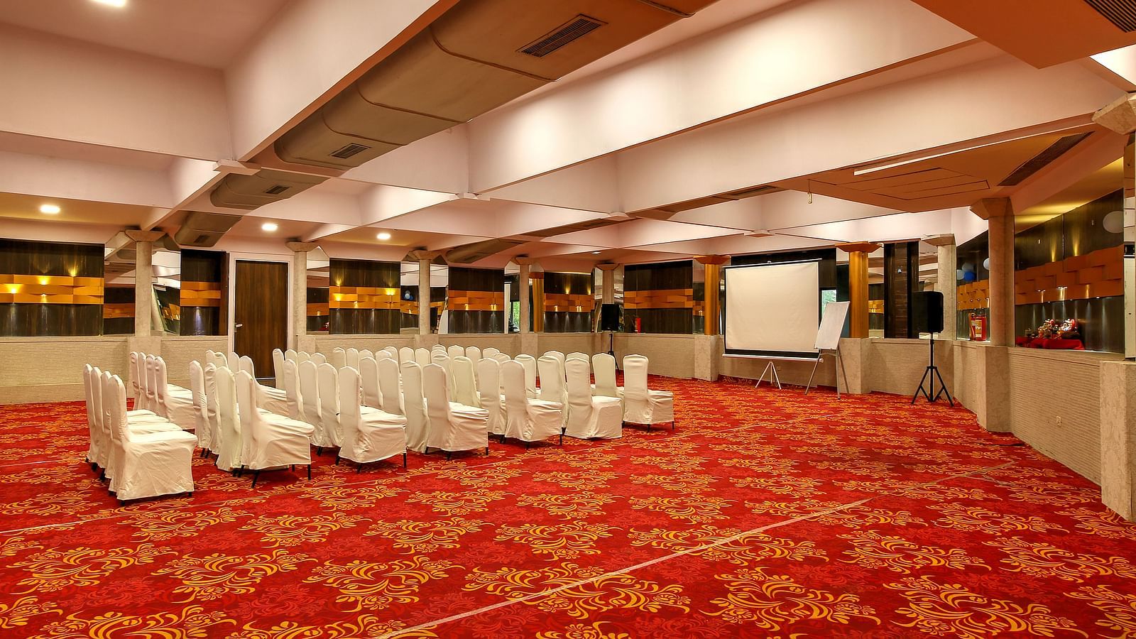 An event hall with red carpet, arranged chairs facing a presentation screen, and a mirrored ceiling.