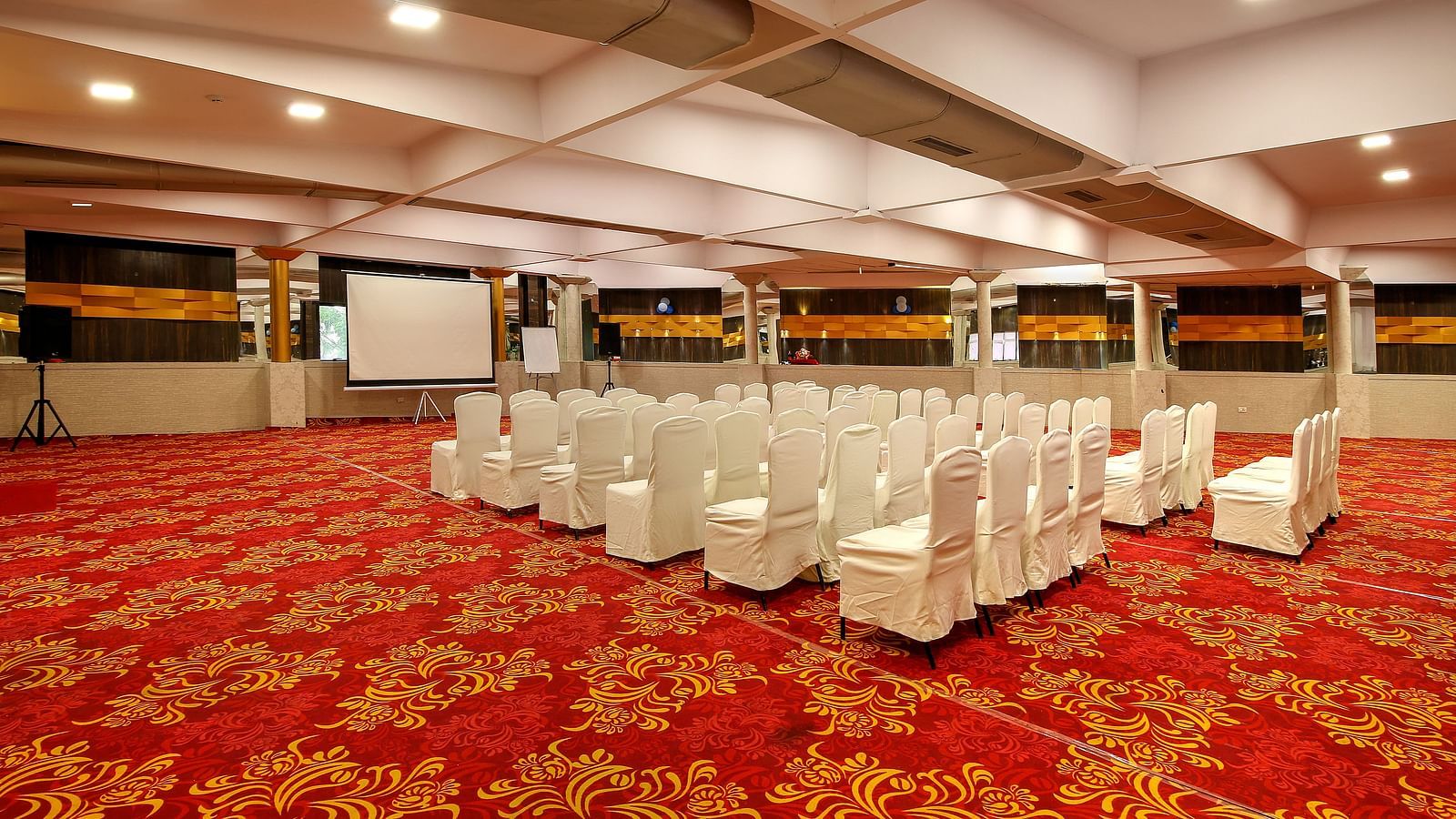 A spacious banquet hall with a patterned red carpet, rows of white-covered chairs facing a projector screen, and a reflective ceiling creating an elegant atmosphere.