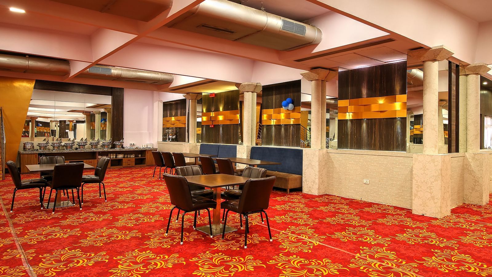 A banquet area with a red patterned carpet, dining tables with chairs, and a bar with blue and gold accents under warm lighting.