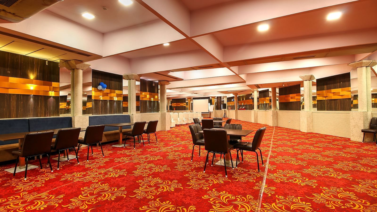 A banquet hall with a red patterned carpet, dining tables with chairs, and a bar with blue and gold accents under warm lighting.