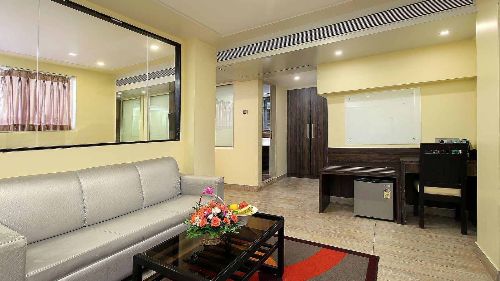 A hotel suite's living area with a grey sofa, a glass coffee table with a fruit basket, and a mirror on the wall reflecting the room. A wooden work desk with a chair and a minibar fridge is seen in the background.