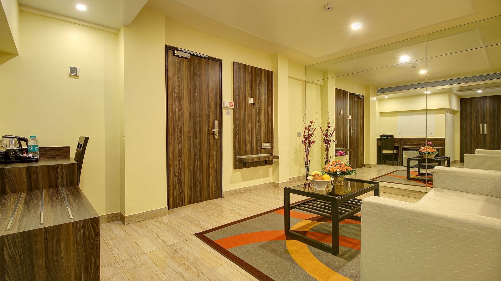 A bright hotel living area with glossy floors, a wooden shelf with a kettle, and water bottles.