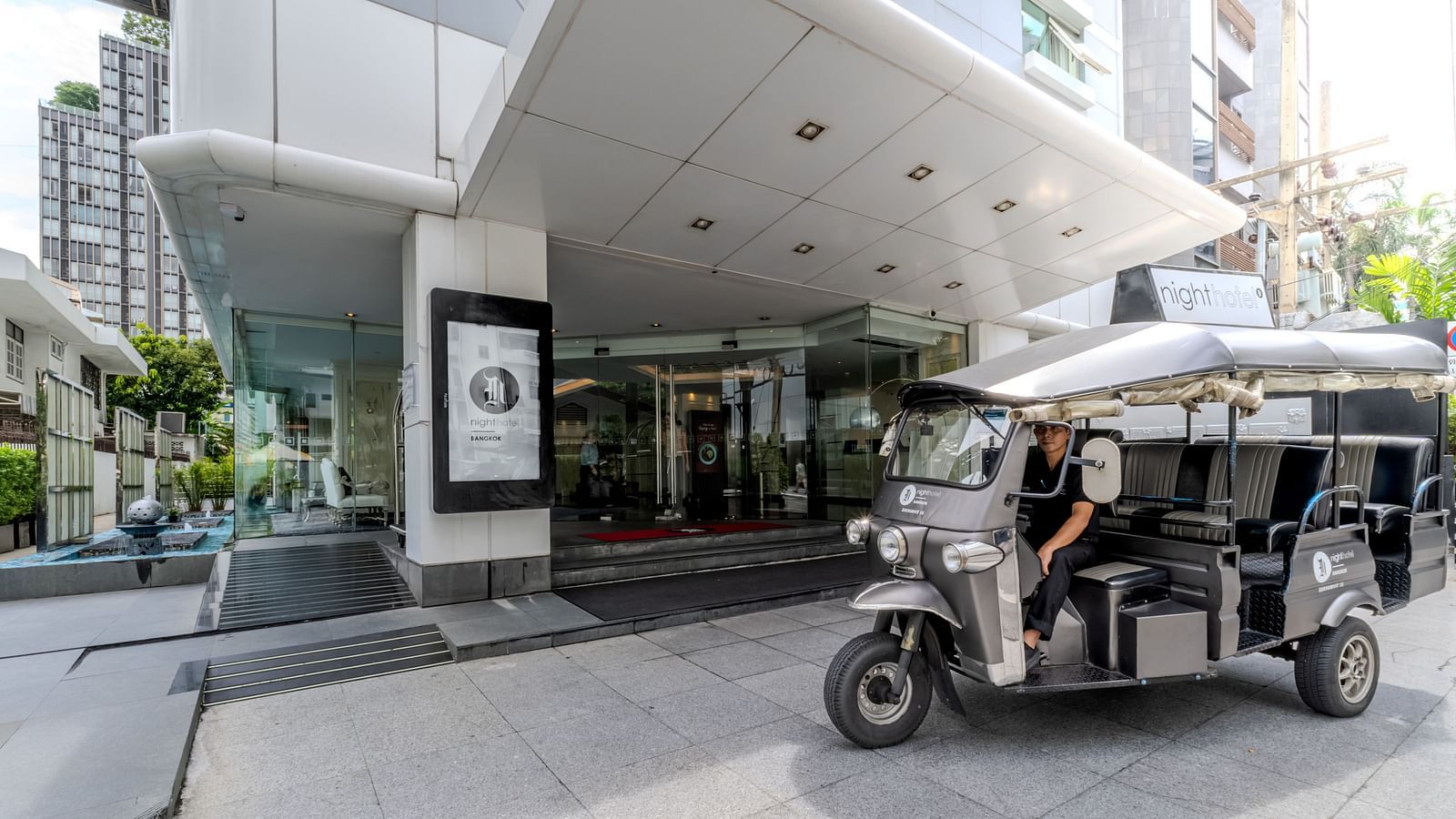 A tuk tuk parked in front of the entrance of Night Hotel Bangkok during the day.