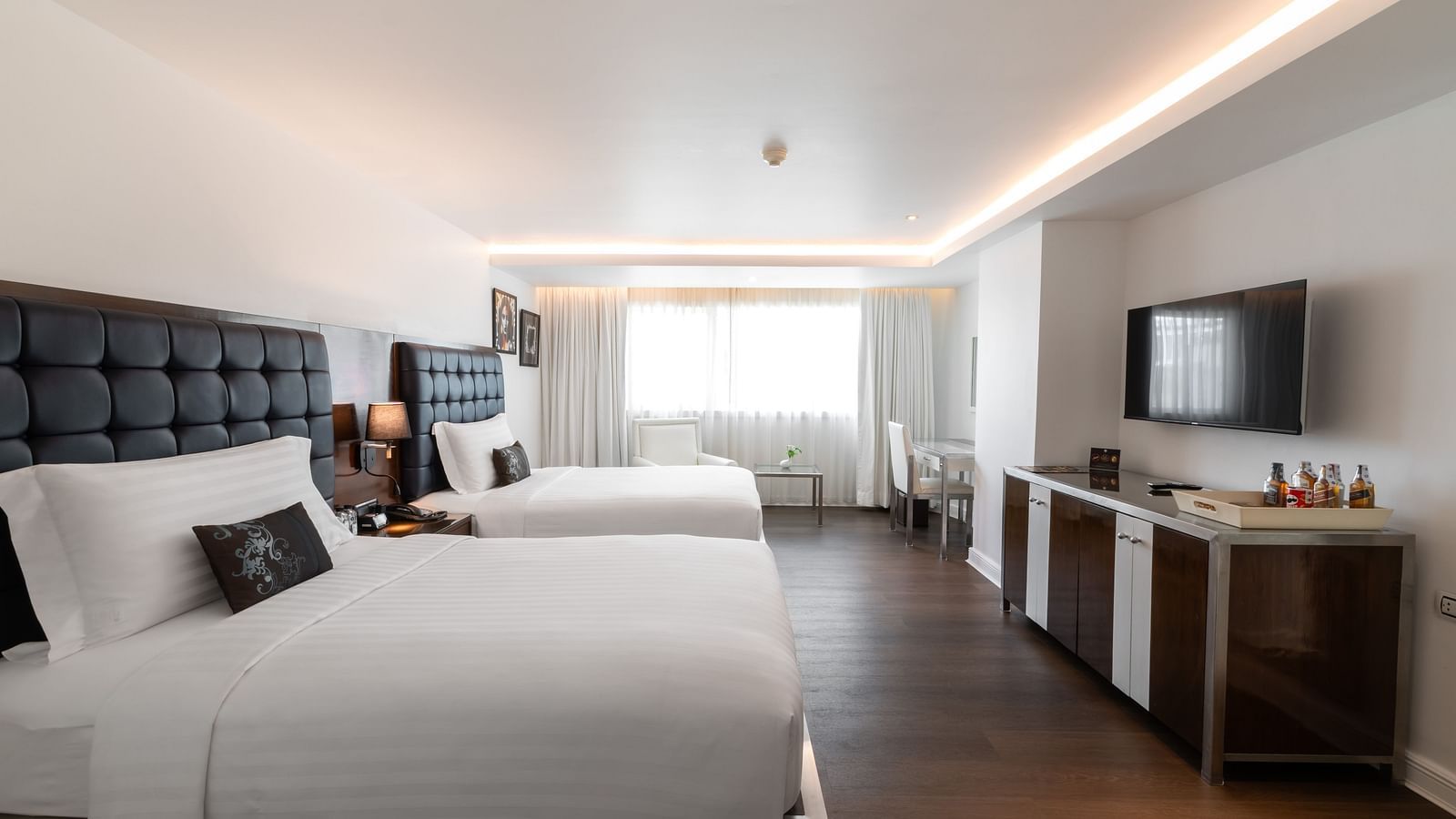 Modern hotel room with two large beds, white linens, dark headboards, a flat-screen TV, minibar, and a window letting in natural light at the far end of the room at Night Hotel Bangkok.