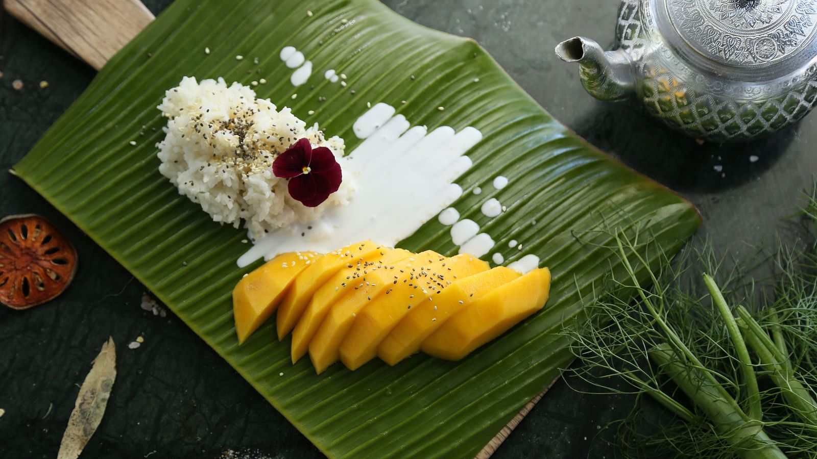 A top view of Mango Sticky Rice served on a banana leaf with a tea pot next to it.