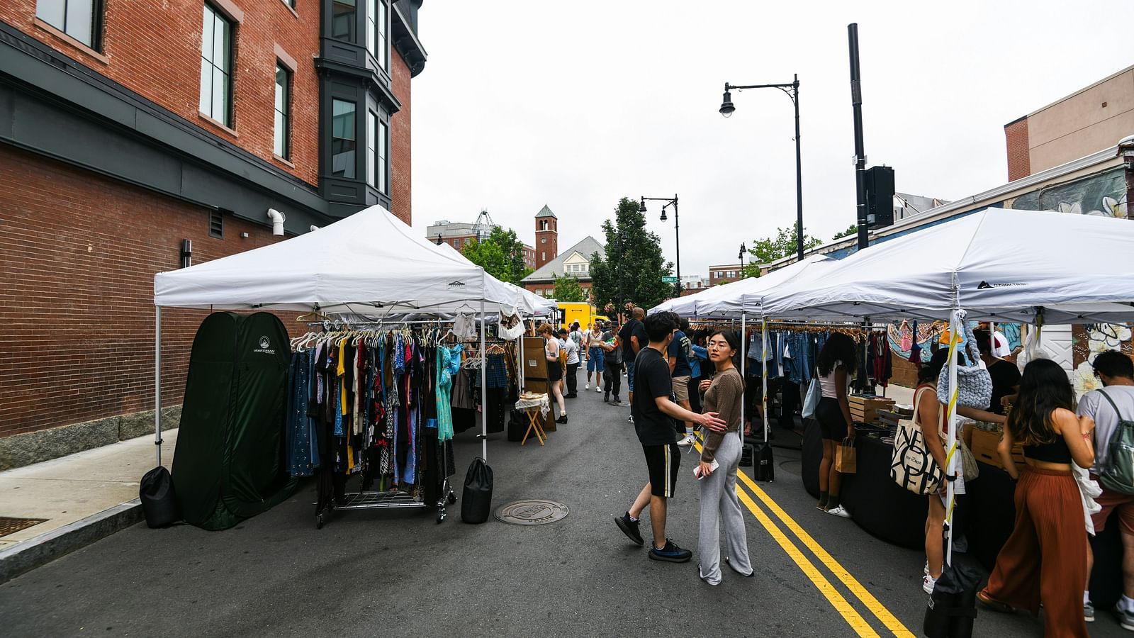 Many shops on the street selling clothes with people looking at them.