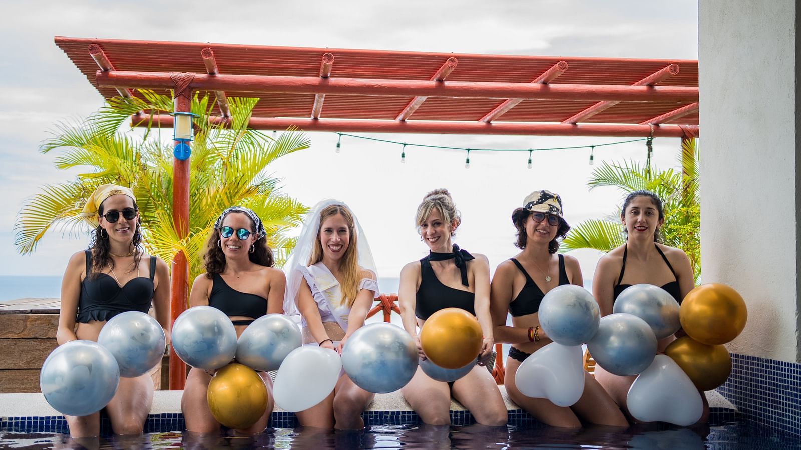 a group of women sitting next to a pool holding silver and gold balloons