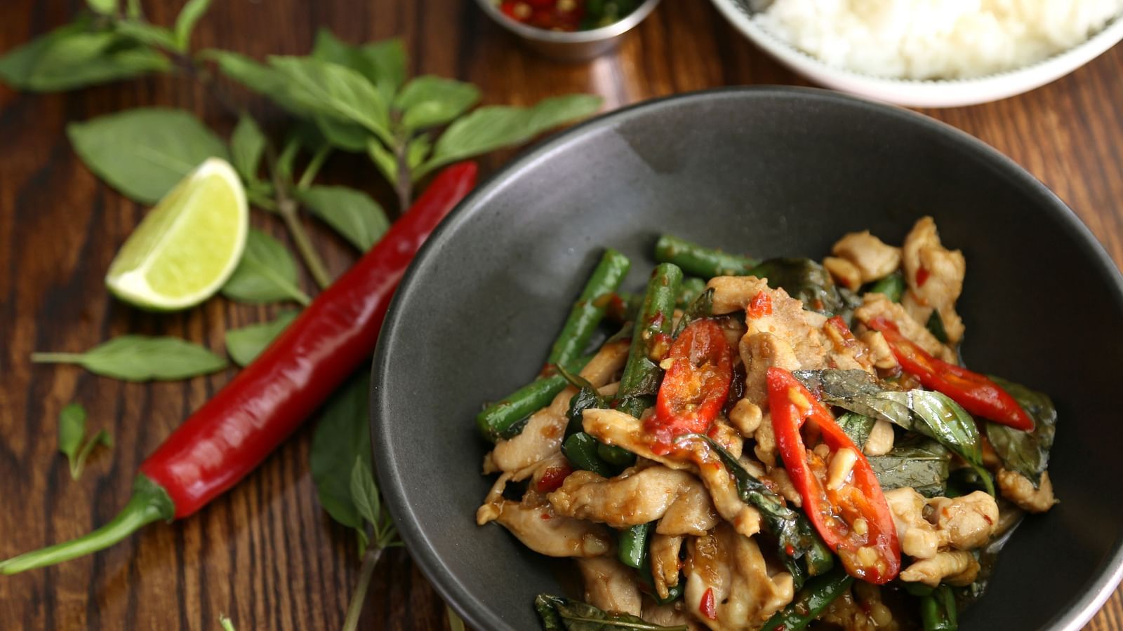 A top view of Stir-Fried Holy Basil kept in a black bowl with a dry chilli and a slice of lime kept next to it.