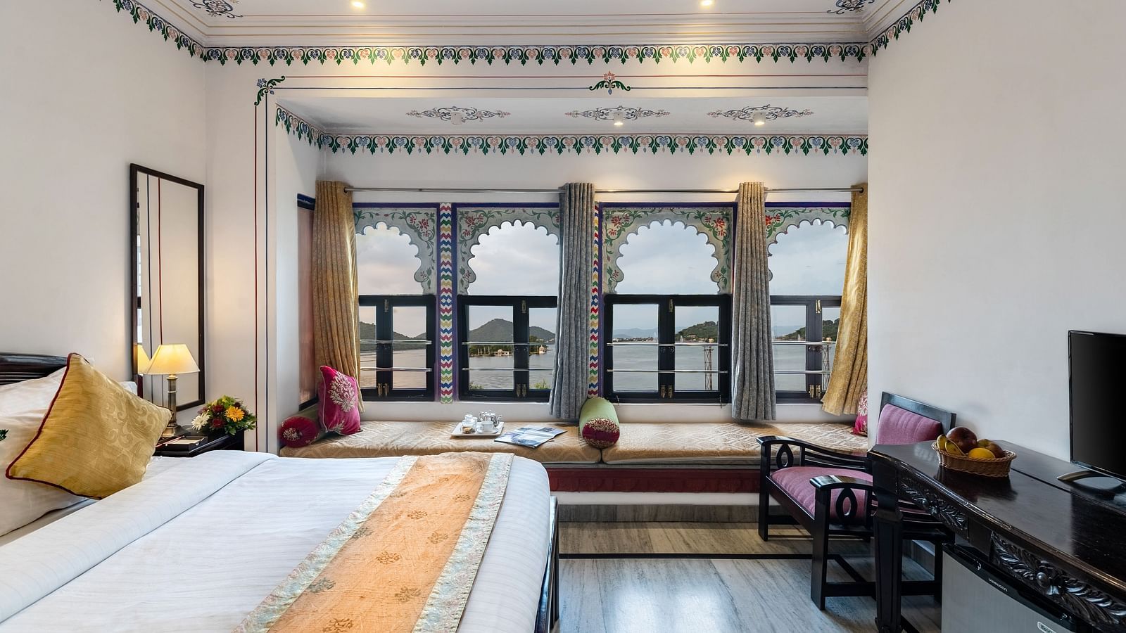 A side view of the bedroom featuring a spacious bed, a wooden table under the wall-mounted television, and bay window seating with curtains pulled to the side at Panna Vilas Palace