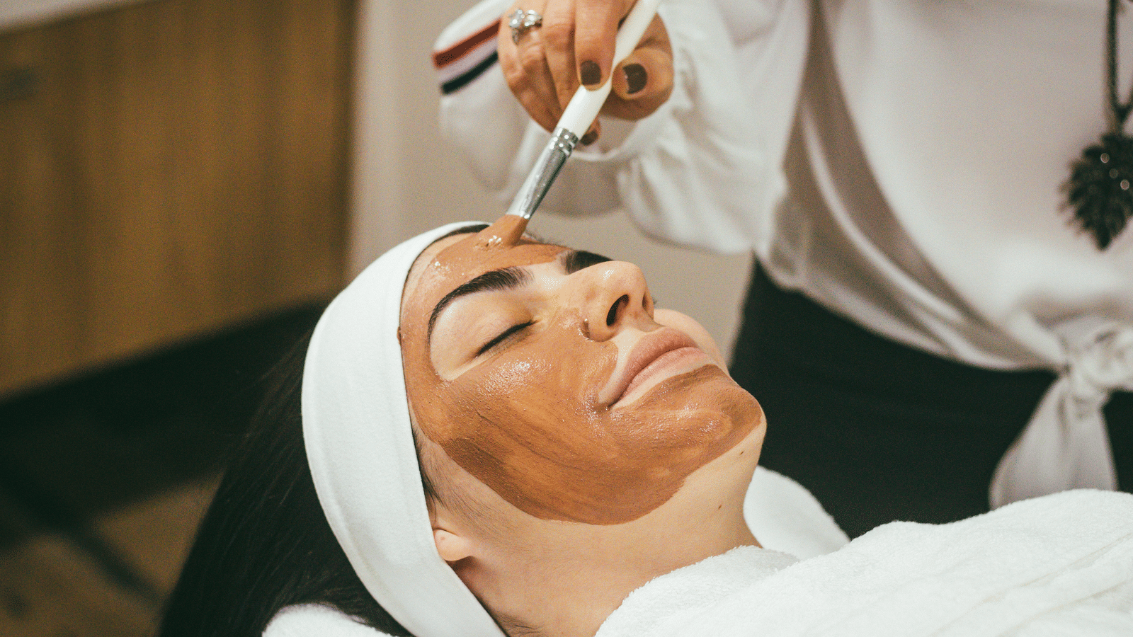 A person uses a brush to apply a clay face mask to a woman's forehead during a professional skincare treatment.
