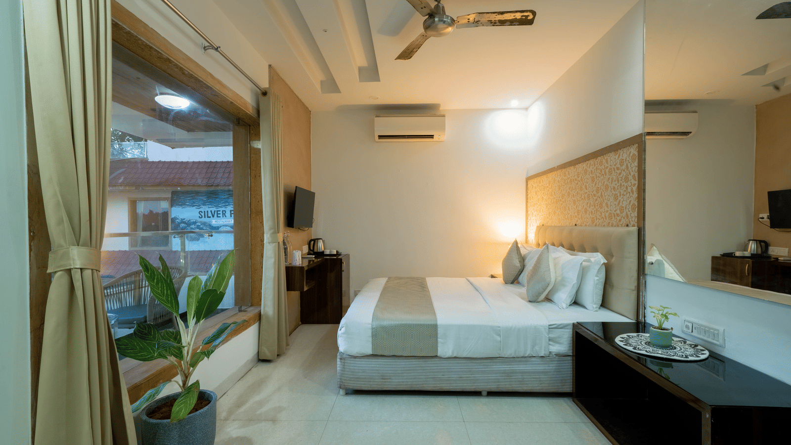 A brightly lit, modern hotel bedroom at Perfectstayz Premium Goa Continental with a large white bed, air-conditioning, an indoor plant kept beside the huge glass window, and light-coloured furnishings.