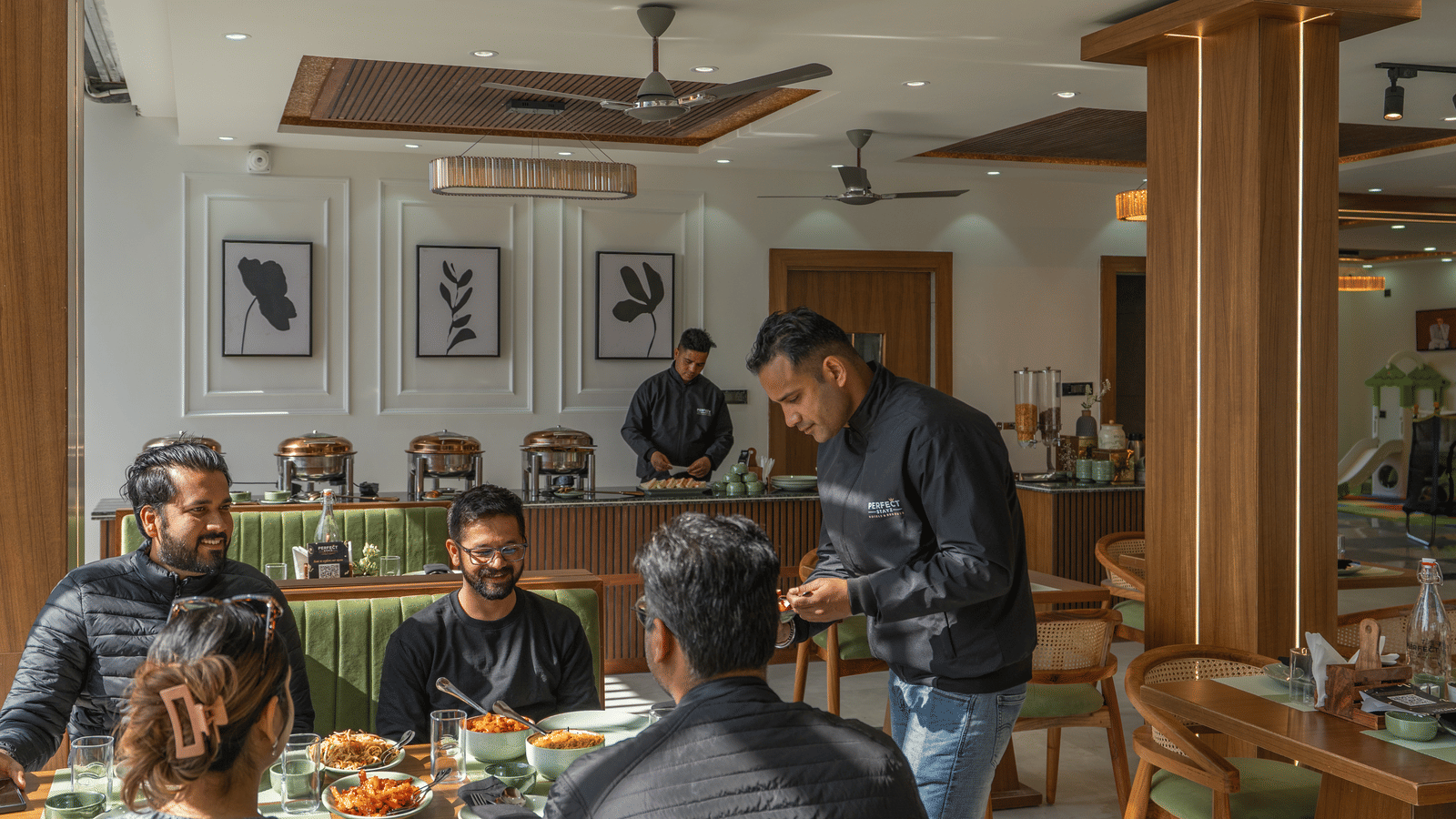4 people sitting with 2 office staff, one serving, and another standing by the reception, at a dining table inside Perfectstayz Premium Kapilaz, Bhimtal.