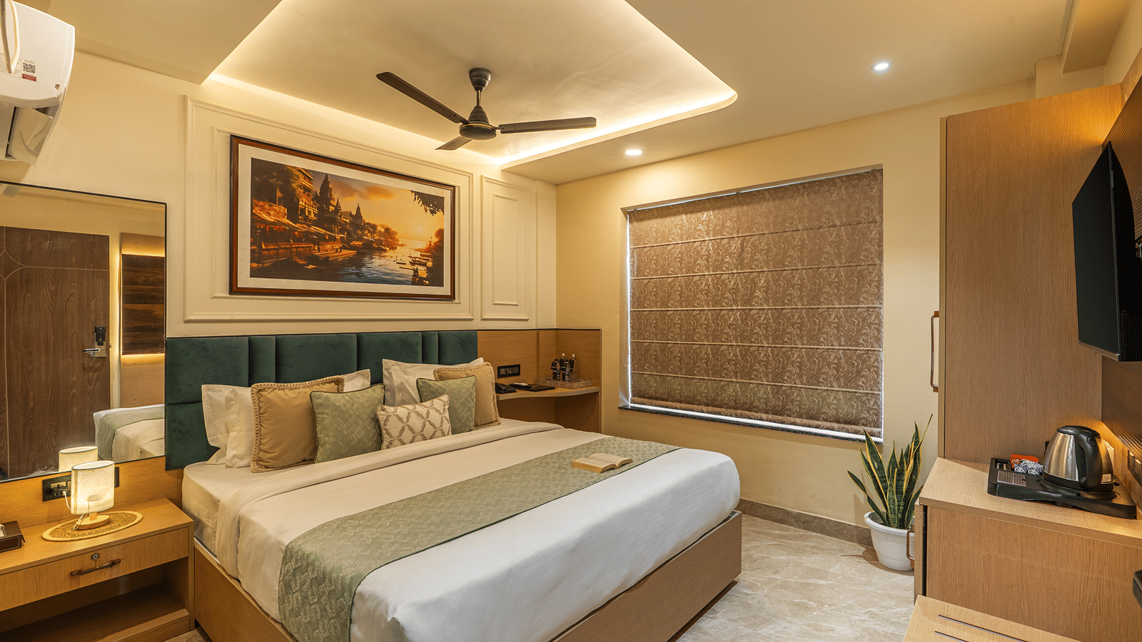 Deluxe Room at Perfectstayz Premium Lavender, Varanasi with a double bed, teal headboard, a ghat painting, cove-lit ceiling, and a patterned blind drawn down.