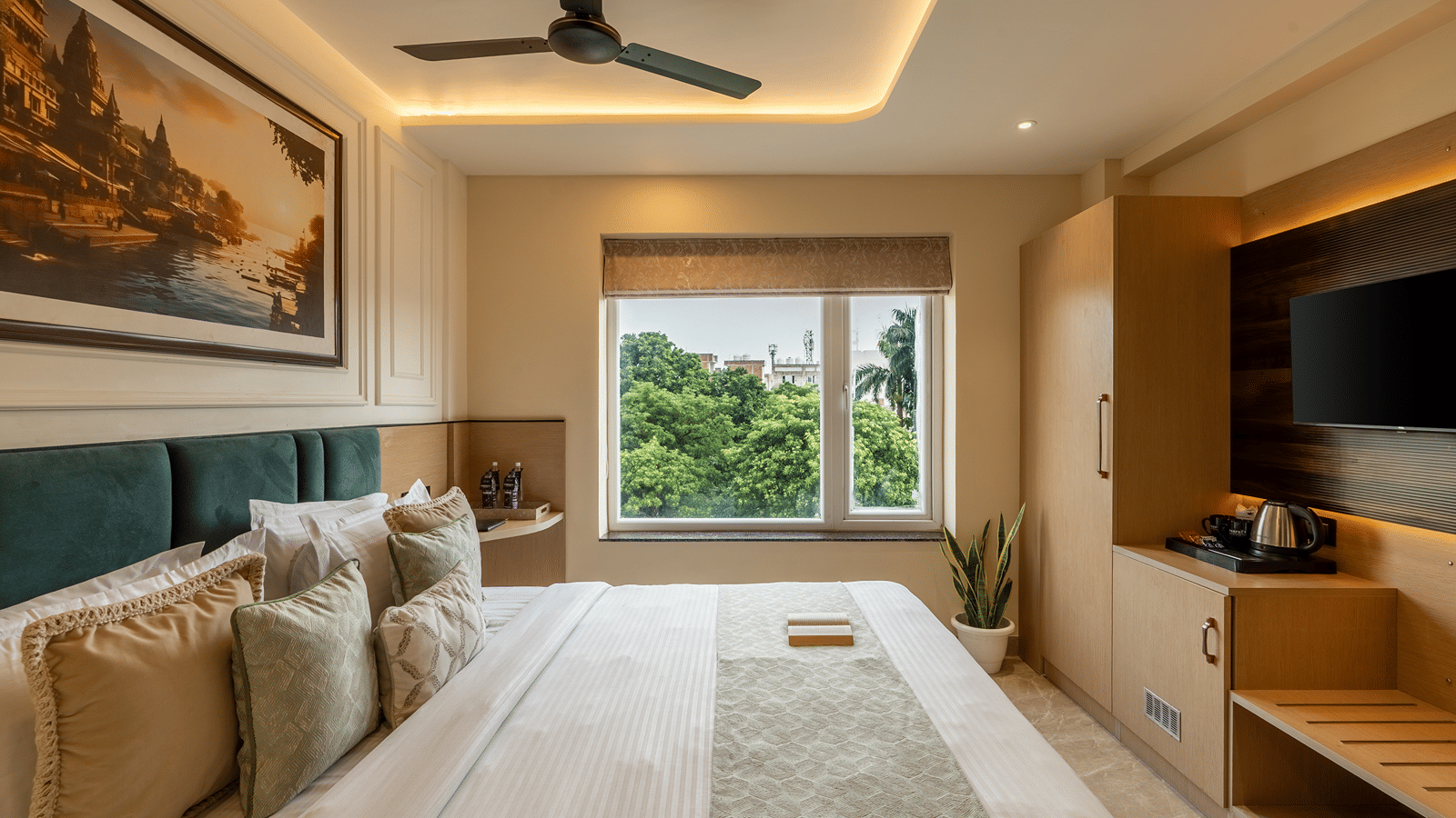 Deluxe Room at Perfectstayz Premium Lavender, Varanasi with a double bed, teal padded headboard, a ghat painting, wall TV, kettle, and a window with tree views.