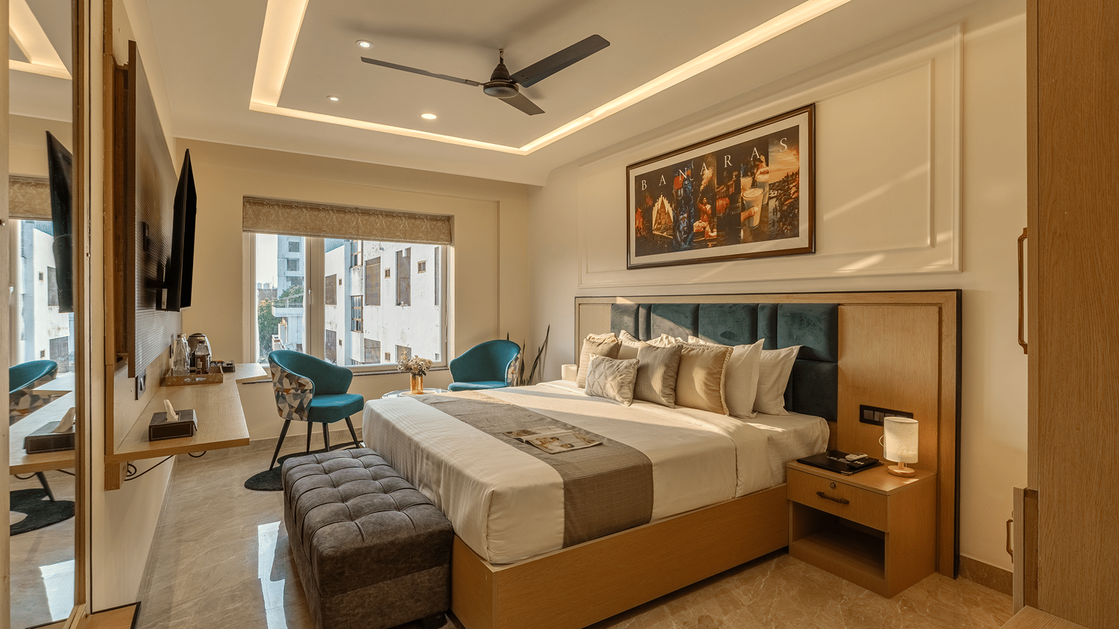 Wide view of the Executive Room at Perfectstayz Premium Lavender, Varanasi with a king bed, grey bench, 2 blue armchairs, wall TV, wardrobe, and a Banaras poster.