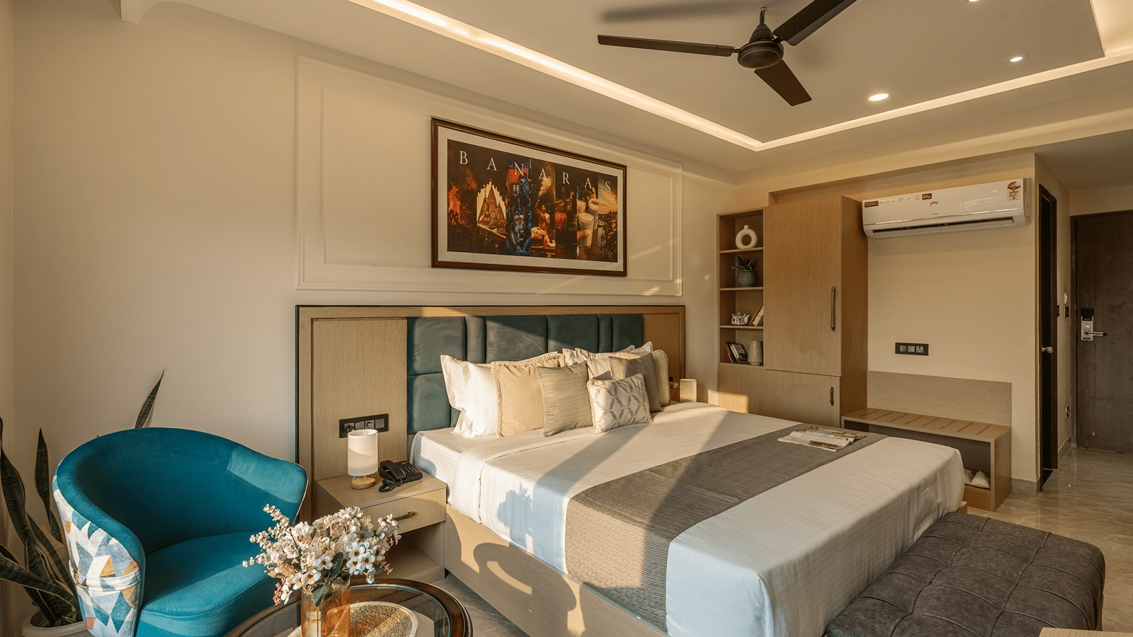 Executive Room at Perfectstayz Premium Lavender, Varanasi with a double bed, teal headboard, a blue armchair, a round glass table with flowers, and a Banaras poster.
