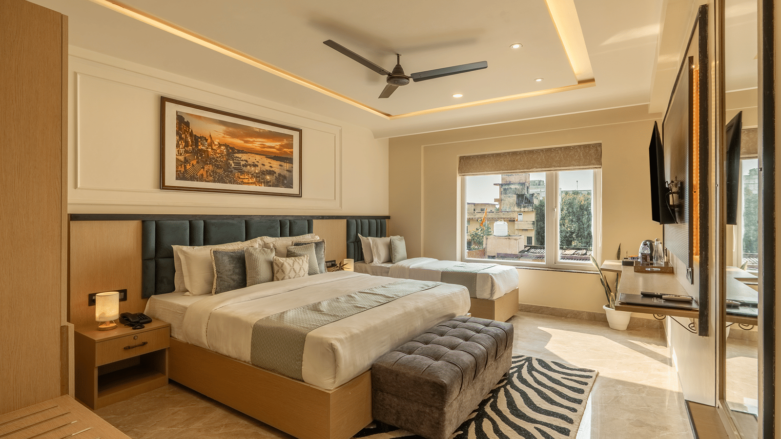 Family Triple Room at Perfectstayz Premium Lavender, Varanasi with a double bed and a single bed by a city view window, teal headboards, a grey bench, and a wall TV.