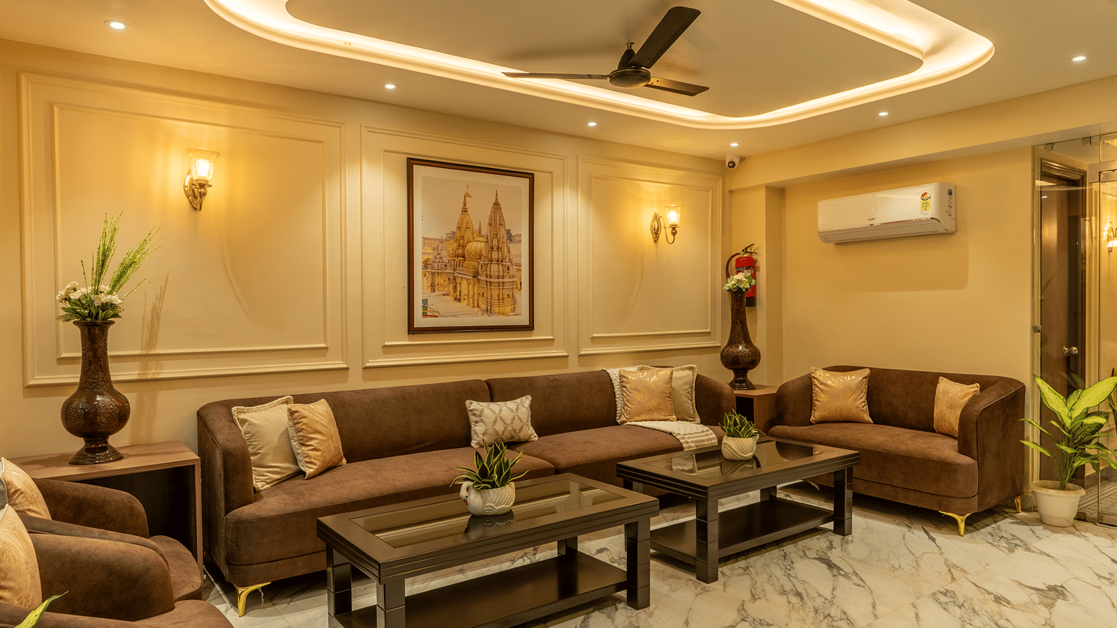Lobby lounge at Perfectstayz Premium Lavender, Varanasi with brown sofas, gold cushions, 2 dark wood coffee tables, a framed temple print, and marble flooring.