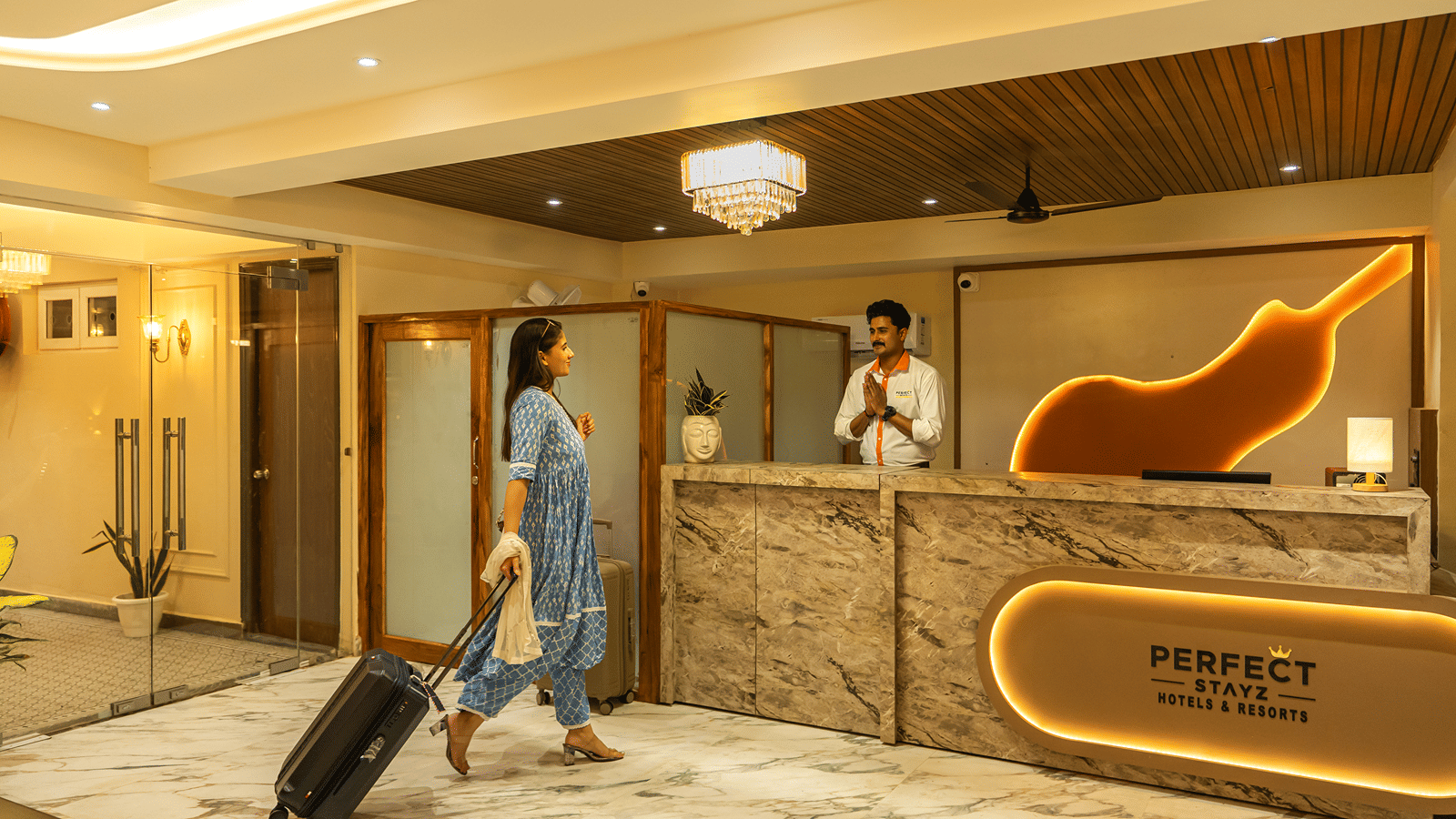 Guest with a suitcase arriving at the reception of Perfectstayz Premium Lavender, Varanasi, greeted by a staff member behind a marble desk with a backlit logo.