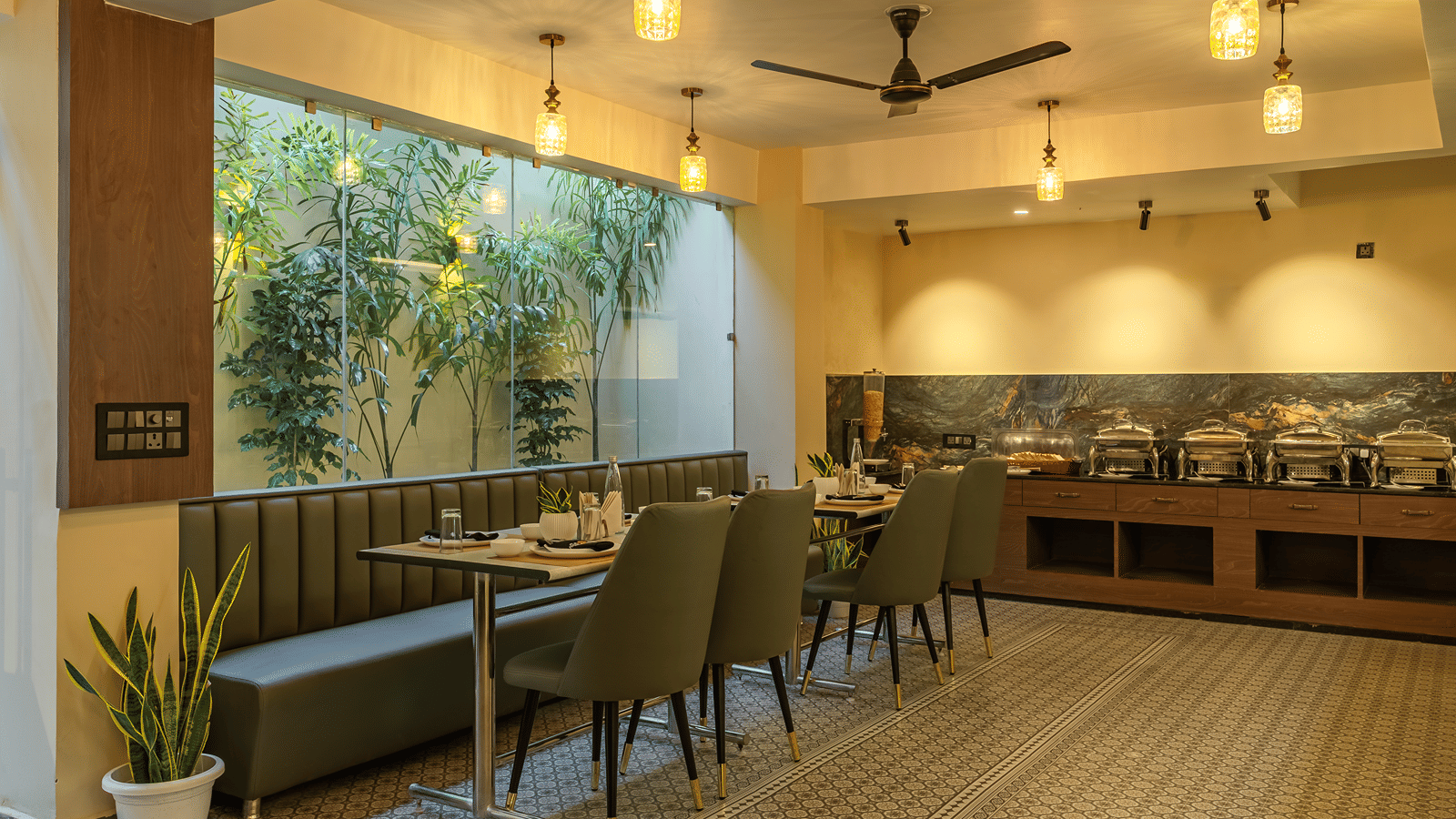 Restaurant at Perfectstayz Premium Lavender, Varanasi with a buffet counter of 4 chafing dishes, green banquette seating, pendant lamps, and a planted glass wall.