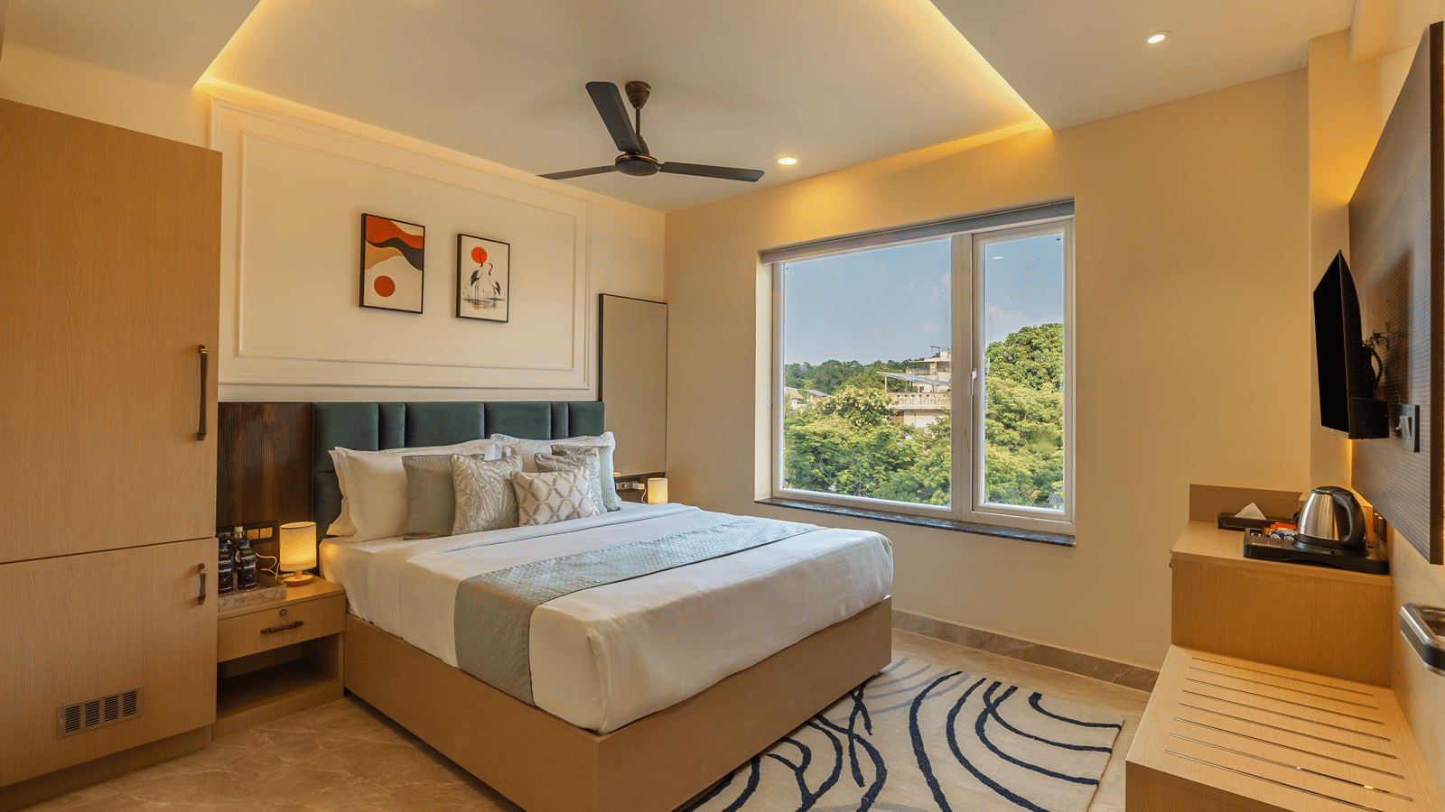 Super Deluxe Room at Perfectstayz Premium Lavender, Varanasi with a double bed, teal headboard, 2 abstract prints, a wavy blue rug, and a window with tree views.