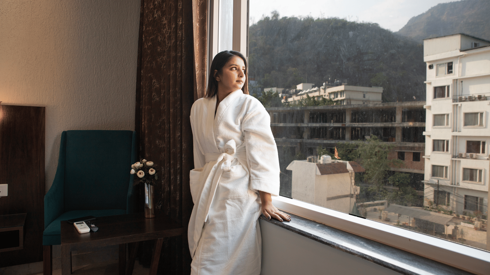 A woman in a bathrobe at Perfectstayz Premium Laxmi Heritage stands by a large window, looking out at buildings and forested hillsides.