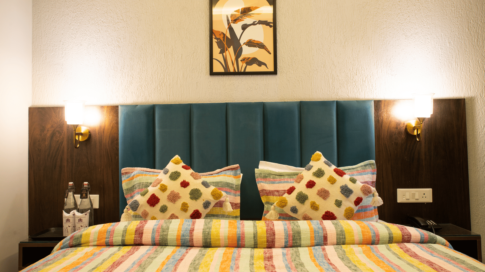 A bedroom at Perfectstayz Premium Laxmi Heritage with a king size bed, 2 patterned throw pillows, 2 bedside tables with water bottles, and 2 wall-mounted lamps.