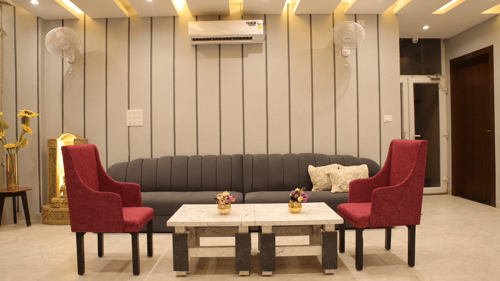 A lobby area at Perfectstayz Premium Laxmi Heritage with a sofa, 2 armchairs, 2 coffee tables, an A/C, and striped wall panelling with decorative lighting.