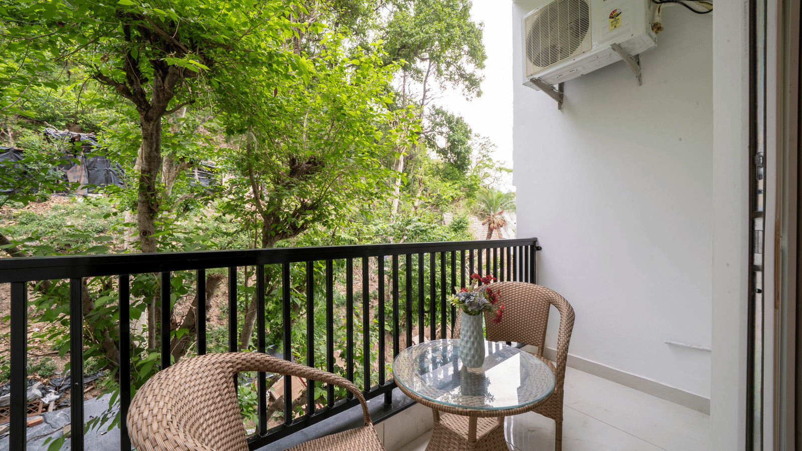 A balcony with an air-conditioning unit, a railing, 2 wicker chairs, a glass table, and trees visible outside the space in Perfectstayz Premium Mystic Falls, Rishikesh.