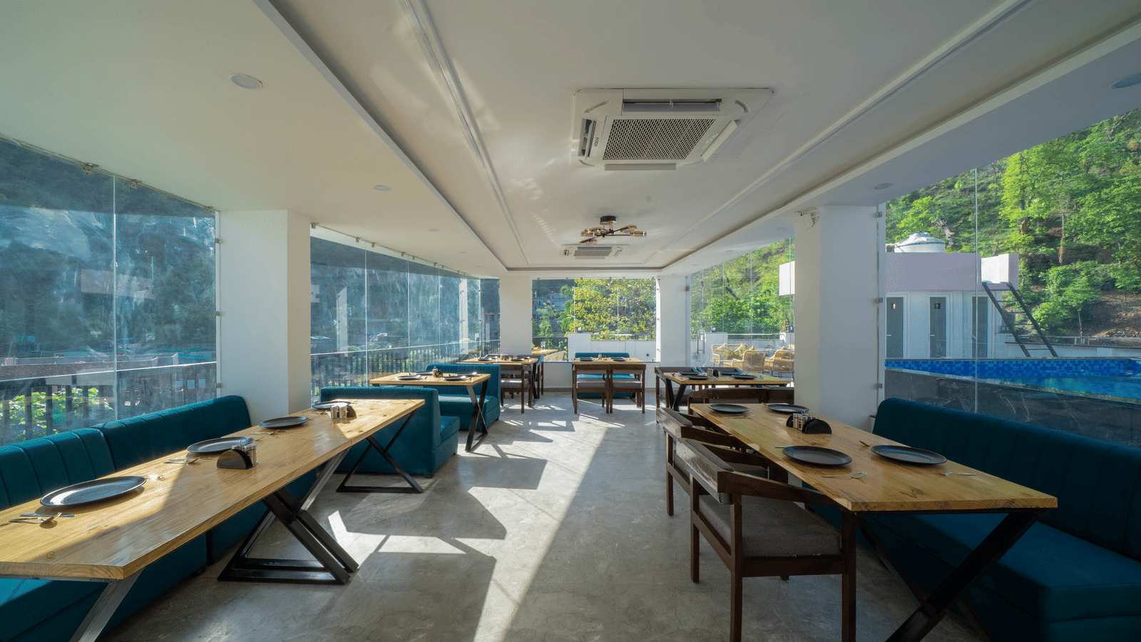 A dining area with tables, benches with cushions, a ceiling-mounted air-conditioning unit, and windows looking out to trees in Perfectstayz Premium Mystic Falls, Rishikesh.
