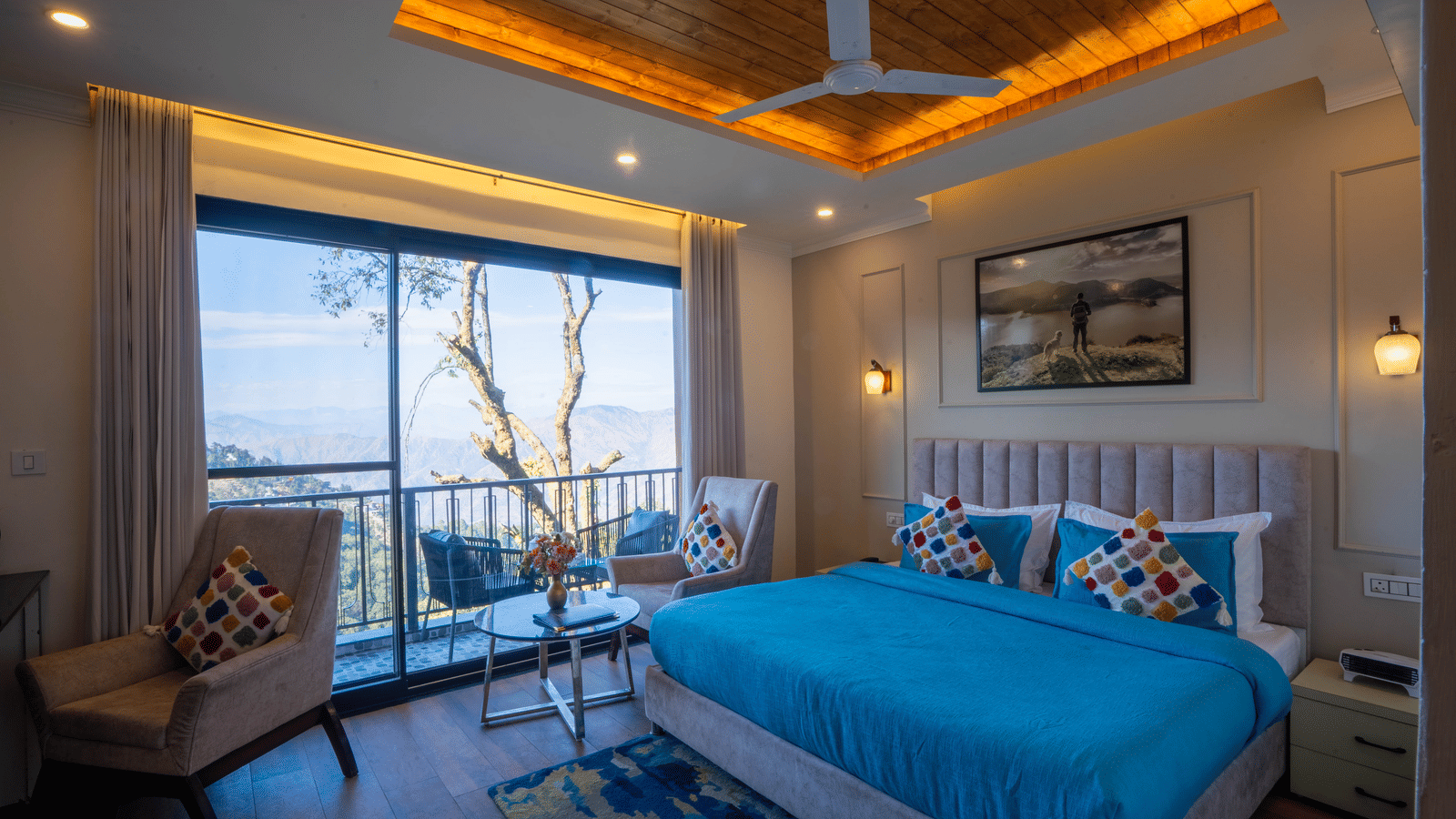 A wide-angle shot of the Executive Room With Balcony & Valley View featuring a double bed with blue sheets and 2 chairs overlooking the valley from the balcony at Perfectstayz Premium Westend Mall Road, Mussoorie.