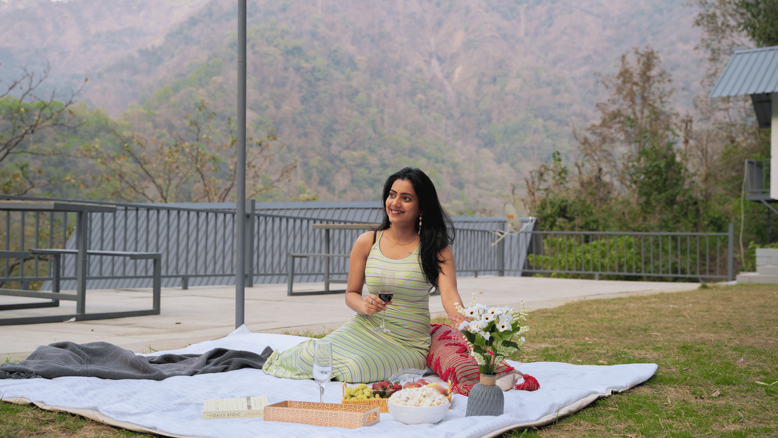 A person sits on a picnic blanket on the grass at Perfect Stayz The Jungle Resort, surrounded by food and flowers.