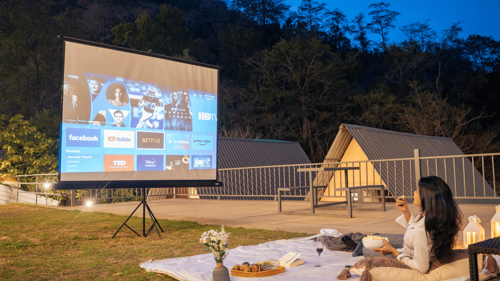 A person sits on a picnic blanket watching a film projected onto a screen at Perfect Stayz The Jungle Resort.