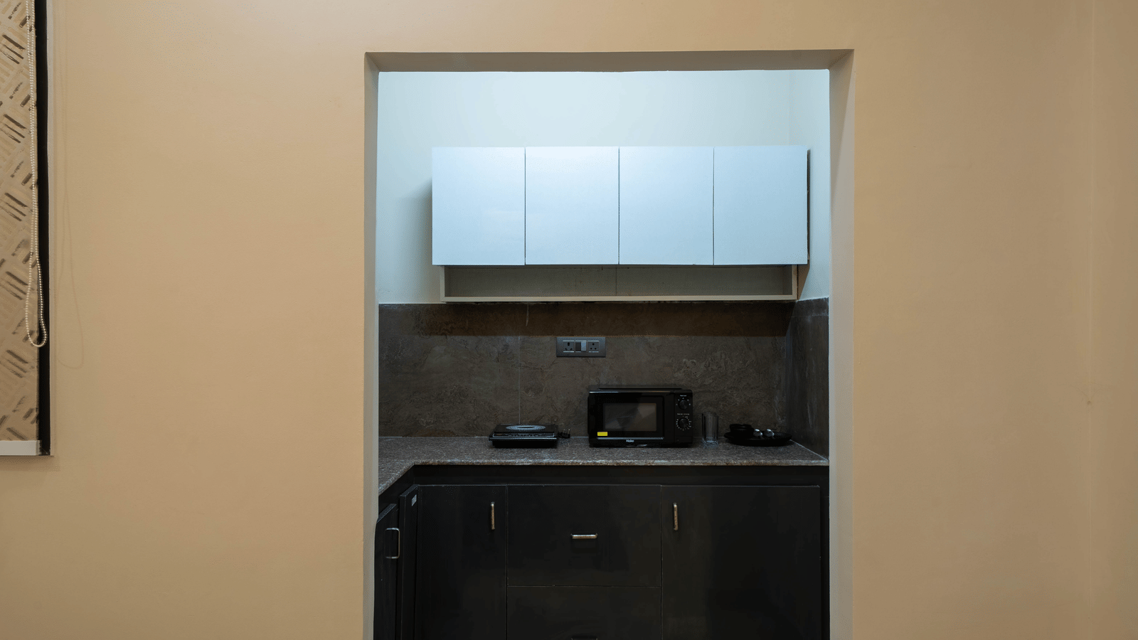 A close view of a built-in kitchenette area with white upper cabinets, a microwave, and dark grey storage set into the wall - Perfectstayz Value Aalaya Suites, Haridwar