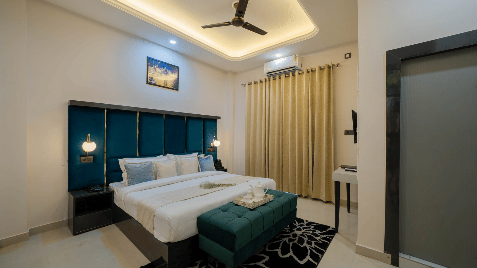 Interior view of a modern bedroom with a bed featuring a teal tufted headboard, white linens, a ceiling fan, and a dark green velvet bench - Perfectstayz Value Aalaya Suites, Haridwar