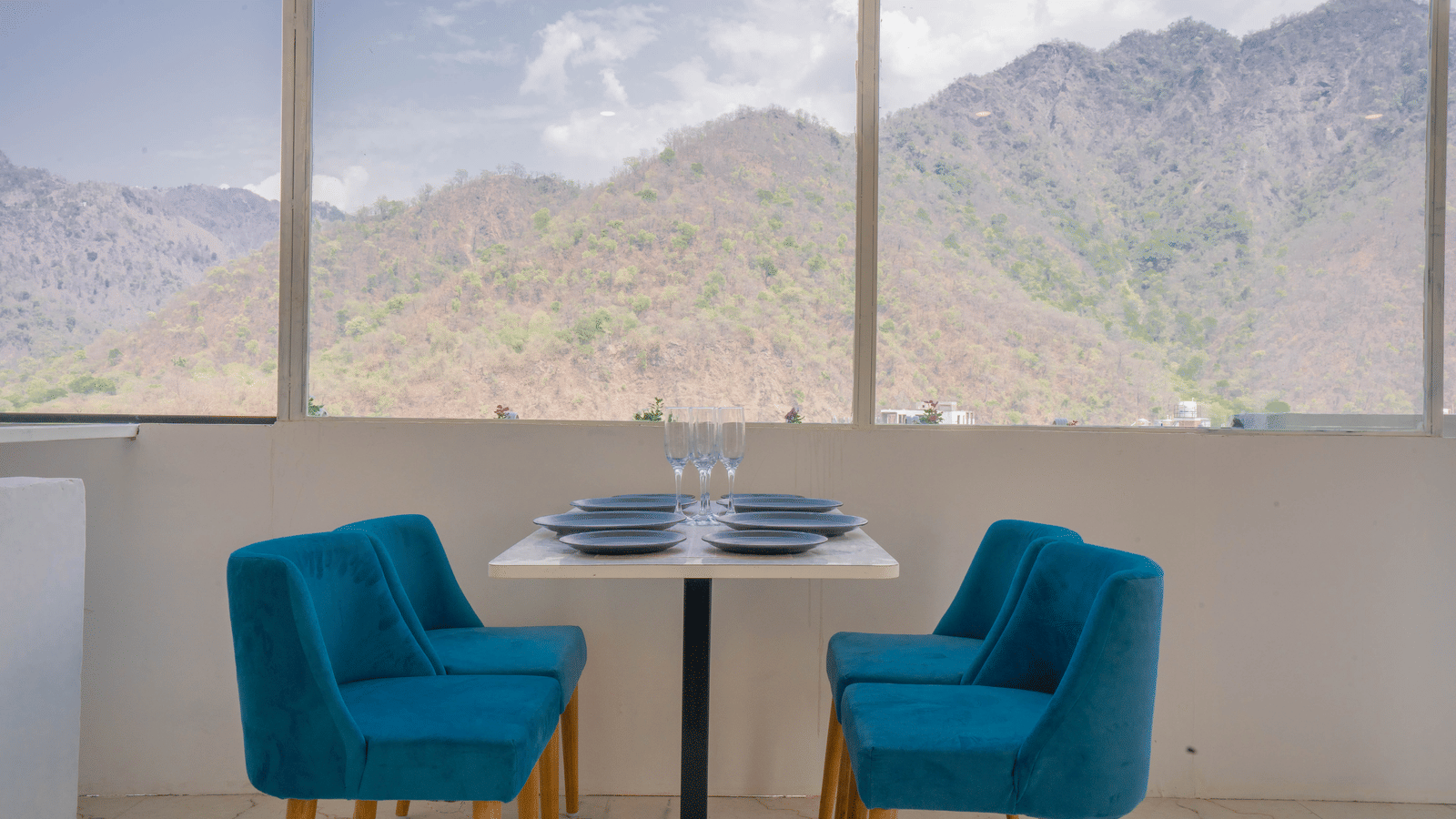 A dining area at Perfectstayz Value Alpine Retreat, Rishikesh with large windows offering beautiful views of the surrounding hills.