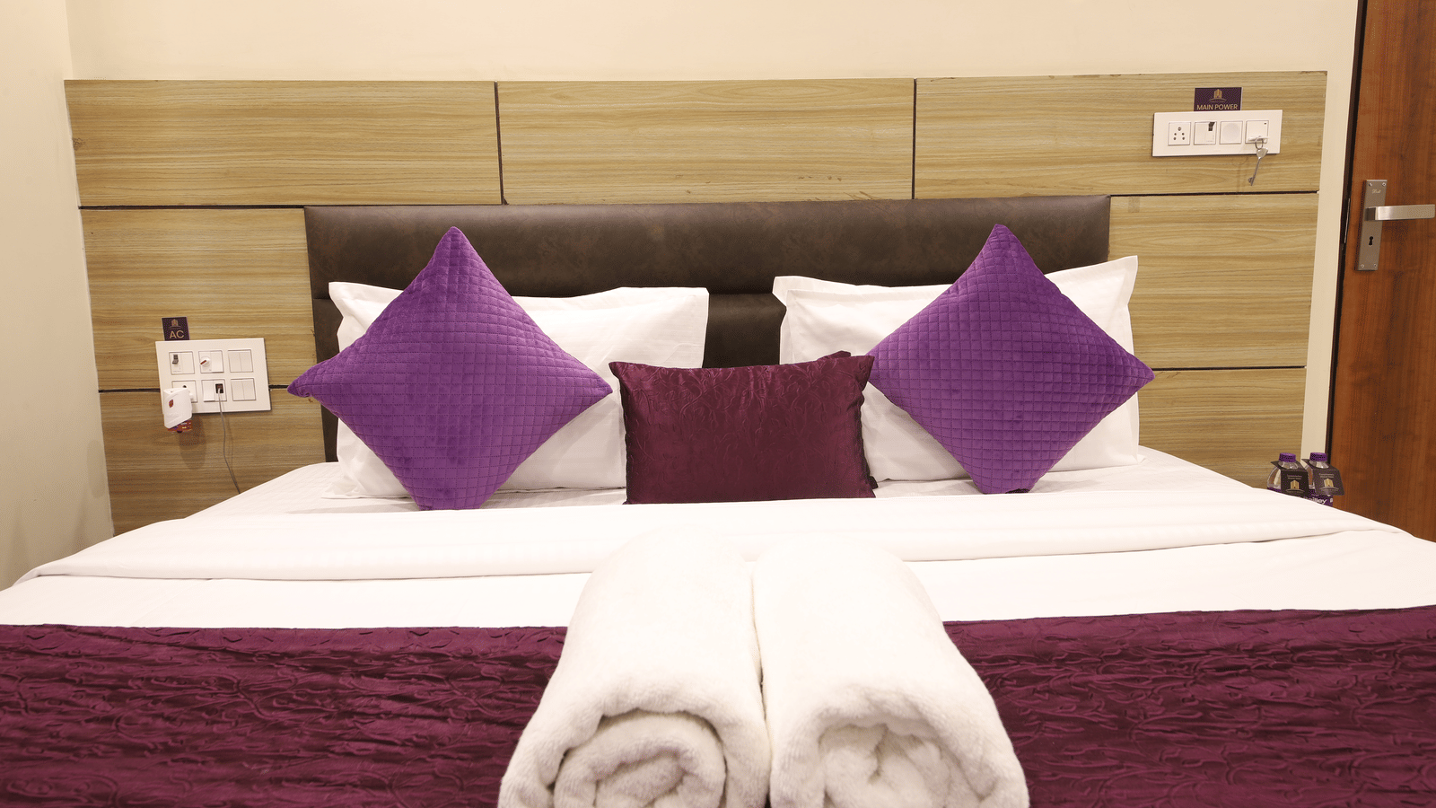 Interior view of a bed with white and purple bedding and rolled towels at Perfectstayz Value Dwarkesh, Haridwar.